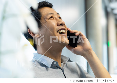Talking on smartphone, asian man smiling and enjoying conversation in office setting Talking on smartphone, asian man smiling and enjoying conversation in office setting 120615897