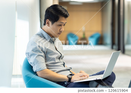 Working on laptop, asian man focusing on business tasks in modern office setting 120615904