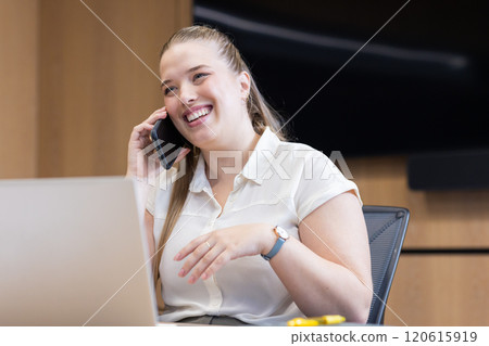 Talking on smartphone, woman smiling while working on laptop in office Talking on smartphone, woman smiling while working on laptop in office 120615919
