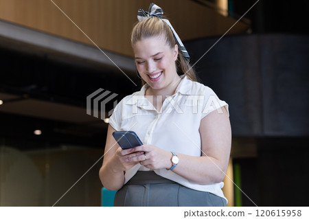 Smiling woman using smartphone in modern office building, wearing business attire 120615958