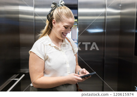 At office, Using smartphone, woman smiling and texting in elevator, business attire At office, Using smartphone, woman smiling and texting in elevator, business attire 120615974