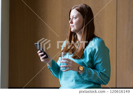 At office, Holding smartphone and glass of water, woman in blue sweater looking away 120616149