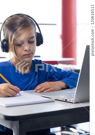In school, boy wearing headphones writing in notebook and using laptop In school, boy wearing headphones writing in notebook and using laptop 120616511