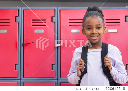 Smiling african american girl with backpack standing in front of red lockers at school, copy space 120616548