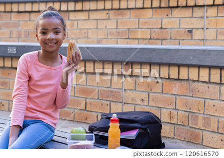 Smiling african american girl eating sandwich during school lunch break with backpack and juice Smiling african american girl eating sandwich during school lunch break with backpack and juice 120616570