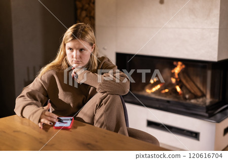 A young woman holding a smartphone, looking reflective while sitting at a wooden table near a warm fireplace in a cozy, softly lit room. A young woman holding a smartphone, looking reflective while sitting at a wooden table near a warm fireplace in a cozy, softly lit room. 120616704