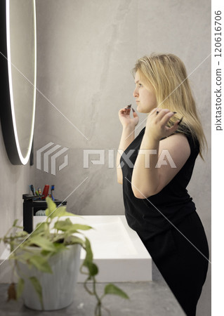 A young woman with blonde hair adjusting her appearance in a modern bathroom, looking at her reflection in a round illuminated mirror. 120616706
