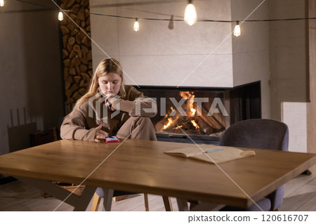 Young woman using a smartphone while sitting at a wooden table in a cozy room with a glowing fireplace and soft string lights. 120616707