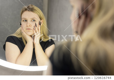 A blonde young woman carefully adjusts her hairstyle while looking into a modern circular mirror with bright illumination in a stylish bathroom. A blonde young woman carefully adjusts her hairstyle while looking into a modern circular mirror with bright illumination in a stylish bathroom. 120616744