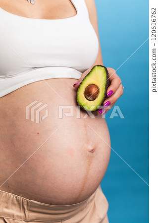 Close-up of a pregnant belly with an avocado held by hand against a blue background 120616762