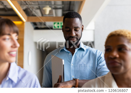 Holding laptop, african american man checking time on watch while standing with colleagues 120616807