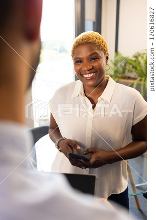 Smiling woman holding smartphone, engaging in conversation with colleague in office Smiling woman holding smartphone, engaging in conversation with colleague in office 120616827
