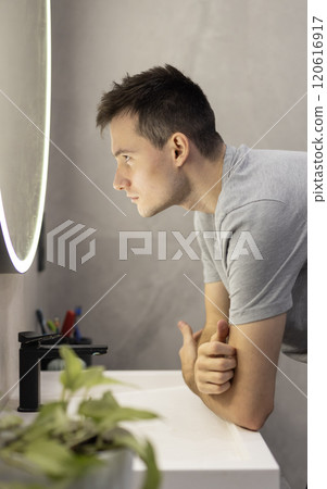 A young man in a grey t-shirt examining his reflection in a modern bathroom mirror with focused attention. A young man in a grey t-shirt examining his reflection in a modern bathroom mirror with focused attention. 120616917
