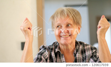 Joyful elderly woman with a triumphant expression, raising her fists in a celebratory gesture indoors. Joyful elderly woman with a triumphant expression, raising her fists in a celebratory gesture indoors. 120617039