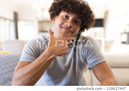 On video call, Smiling man sitting on couch, posing playfully with hand on chin, at home 120617447