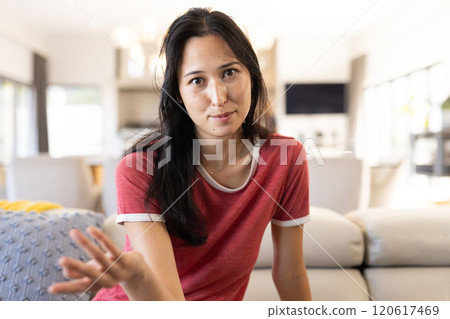 On video call, Woman in casual red shirt gesturing while sitting on couch in living room, at home On video call, Woman in casual red shirt gesturing while sitting on couch in living room, at home 120617469