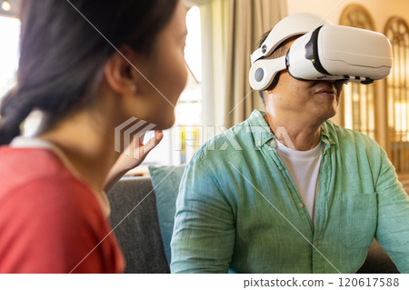 Using VR headset, asian father experiencing virtual reality while woman watching attentively Using VR headset, asian father experiencing virtual reality while woman watching attentively 120617588
