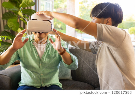 Using VR headset, asian father experiencing virtual reality with assistance in living room 120617593