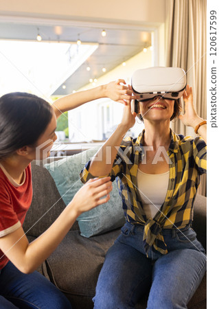 Using VR headset, mother smiling while friend helps adjust it in living room 120617599
