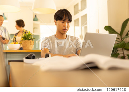 Working on laptop, young man focusing while family in background in kitchen Working on laptop, young man focusing while family in background in kitchen 120617628