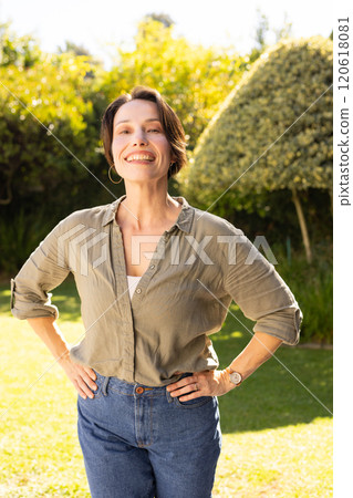 Smiling middle-aged woman standing outdoors with hands on hips, enjoying sunny day 120618081