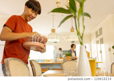 Setting table with plates, smiling asian teenage man in kitchen with family in background Setting table with plates, smiling asian teenage man in kitchen with family in background 120618114