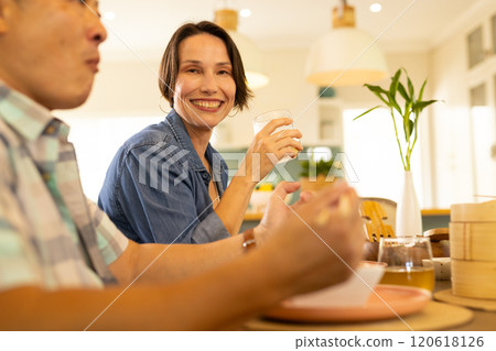 At home, Smiling middle-aged woman enjoying meal with family, holding glass at dining table At home, Smiling middle-aged woman enjoying meal with family, holding glass at dining table 120618126