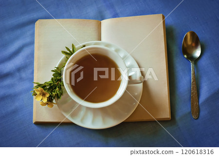 Warm cup of tea placed on an open book with sprig of yellow flowers, set on vibrant blue tablecloth Warm cup of tea placed on an open book with sprig of yellow flowers, set on vibrant blue tablecloth 120618316