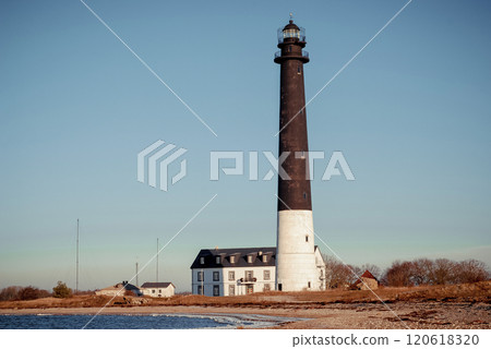 Tall lighthouse on tranquil shore surrounded by smaller buildings, bathed in soft sunlight 120618320