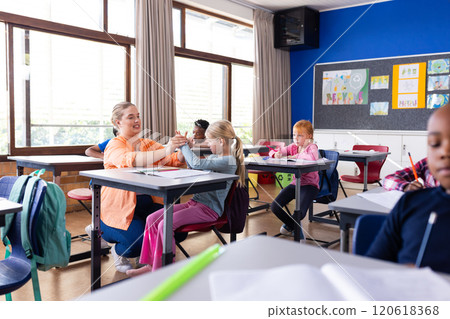 Female teacher assisting young girl with schoolwork in classroom, other diverse students studying 120618368