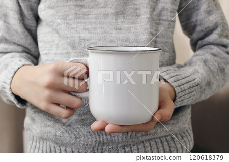 Closeup of hands, fingers holding white cup of tea. Blank cearmic mug mockup. Blurred background, selective focus. Coffee time. Winter still life. Child in grey knitted sweater. Christmas beverage Closeup of hands, fingers holding white cup of tea. Blank cearmic mug mockup. Blurred background, selective focus. Coffee time. Winter still life. Child in grey knitted sweater. Christmas beverage 120618379
