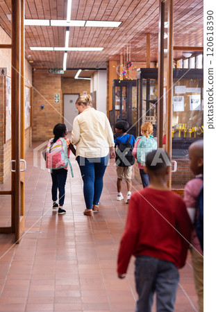 Walking in school hallway, female teacher guiding young diverse students with backpacks Walking in school hallway, female teacher guiding young diverse students with backpacks 120618390