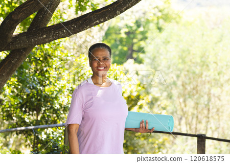 Happy african american senior woman holding exercise mat and smiling in sunny garden, copy space 120618575