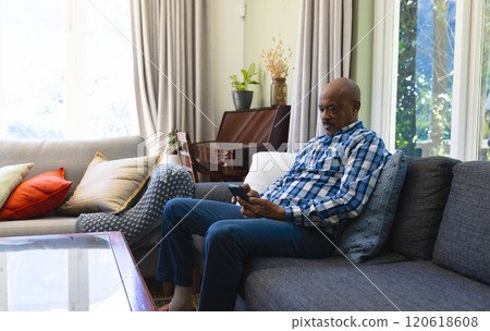 Happy african american senior man sitting on sofa and using smartphone in sunny living room Happy african american senior man sitting on sofa and using smartphone in sunny living room 120618608