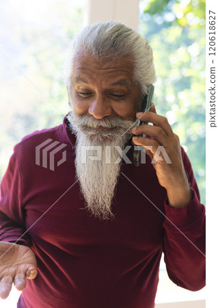 Happy biracial senior man with long beard, using smartphone in sunny living room, copy space 120618627