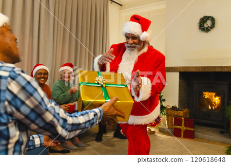 Happy biracial man in santa clothes giving gift to friend next to fireplace at home Happy biracial man in santa clothes giving gift to friend next to fireplace at home 120618648