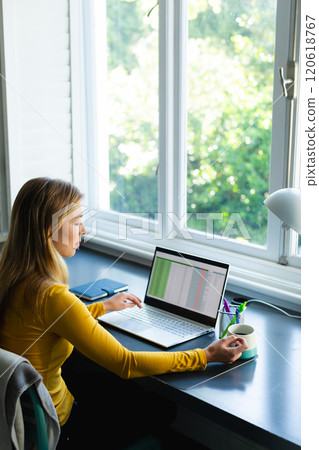 Caucasian woman sitting at desk with coffee, working from home and using laptop in sunny room Caucasian woman sitting at desk with coffee, working from home and using laptop in sunny room 120618767