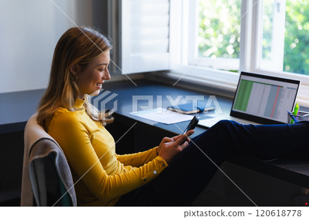 Caucasian woman sitting at desk, working from home, using laptop and smartphone in sunny room 120618778