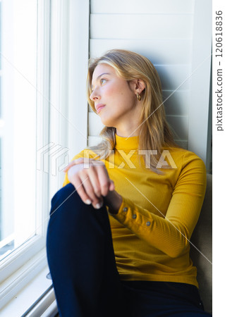 Pensive caucasian woman sitting on windowsill and looking out window in sunny room at home 120618836