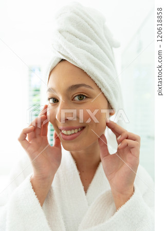Happy caucasian woman in bathrobe looking in mirror, touching face and smiling in sunny bathroom Happy caucasian woman in bathrobe looking in mirror, touching face and smiling in sunny bathroom 120618858