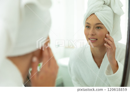 Happy caucasian woman in bathrobe looking in mirror, touching face and smiling in sunny bathroom 120618859