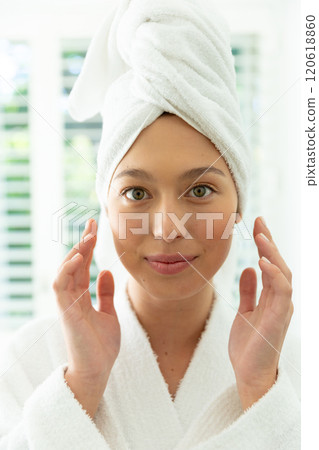 Happy caucasian woman in bathrobe looking in mirror, touching face and smiling in sunny bathroom Happy caucasian woman in bathrobe looking in mirror, touching face and smiling in sunny bathroom 120618860
