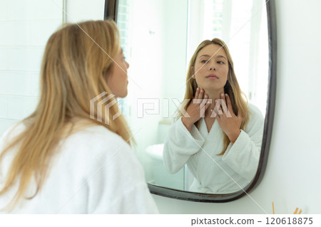 Happy caucasian woman in bathrobe looking in mirror and touching neck in sunny bathroom Happy caucasian woman in bathrobe looking in mirror and touching neck in sunny bathroom 120618875
