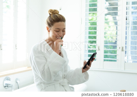 Happy caucasian woman in bathrobe brushing teeth and using smartphone in sunny bathroom Happy caucasian woman in bathrobe brushing teeth and using smartphone in sunny bathroom 120618880