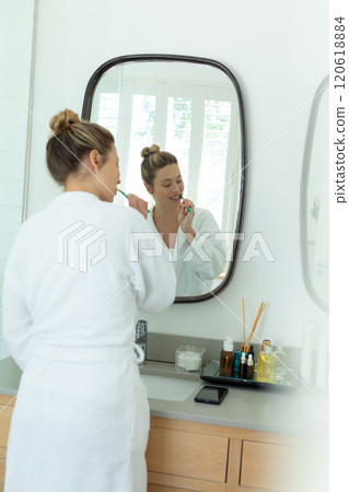 Happy caucasian woman in bathrobe brushing teeth and using smartphone in sunny bathroom Happy caucasian woman in bathrobe brushing teeth and using smartphone in sunny bathroom 120618884