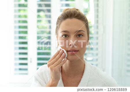 Happy caucasian woman in bathrobe cleaning face with cotton pad in sunny bathroom Happy caucasian woman in bathrobe cleaning face with cotton pad in sunny bathroom 120618889