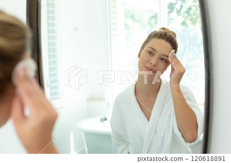 Happy caucasian woman in bathrobe cleaning face with cotton pad in sunny bathroom 120618891