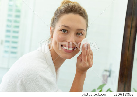 Happy caucasian woman in bathrobe cleaning face with cotton pad and smiling in sunny bathroom Happy caucasian woman in bathrobe cleaning face with cotton pad and smiling in sunny bathroom 120618892