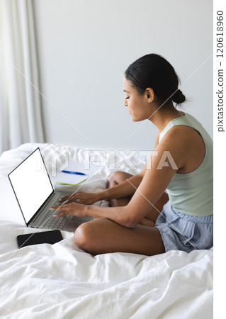 Busy biracial woman with smartphone and notebook using laptop with copy space in bedroom 120618960