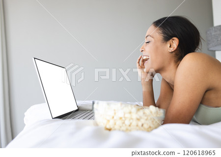 Happy biracial woman with popcorn watching movie on laptop with copy space in bedroom 120618965
